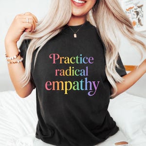 Puede incluir: Camiseta negra con la frase "Practice radical empathy" en un degradado arcoíris. La camiseta es de cuello redondo. La imagen muestra la camiseta en una persona.