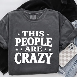 Könnte beinhalten: Dunkelgraues Sweatshirt mit dem weißen Text "THIS PEOPLE ARE CRAZY". Das Bild zeigt gefaltete blaue Jeans und ein Telefon mit karierter Hülle.