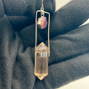 May include: A silver-toned metal pendant featuring a rectangular frame that holds a clear, faceted, hexagonal crystal point at the base and a smaller, purple-hued, irregular gemstone near the top, suspended from a small silver-toned metal loop.