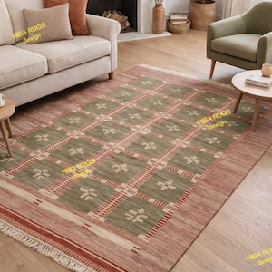 Op de afbeelding: Een rechthoekig vloerkleed met een geometrisch patroon in groen, rood en beige. Het kleed heeft een herhalend vierkant ontwerp met bloemmotieven. De tekst "HIBA RUGS design" is zichtbaar op het kleed.