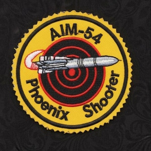 Puede incluir: Parche bordado con un borde circular amarillo y un borde festoneado negro. El parche presenta el texto "AIM-54 Phoenix Shooter" y un diseño de misil sobre un objetivo rojo y negro. El fondo es negro.
