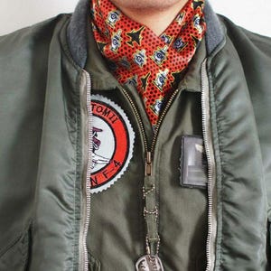 Puede incluir: Chaqueta bomber verde oliva con cuello gris y parche rojo, blanco y negro. La chaqueta tiene cierre de cremallera y una bufanda estampada. Una chapa de identificación plateada cuelga de la cremallera.