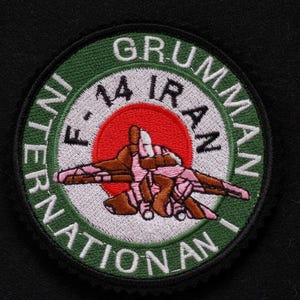 Peut inclure: Écusson brodé circulaire avec un avion de chasse stylisé en marron et rose, sur fond de soleil rouge. Le patch comprend le texte "GRUMMAN F-14 IRAN INTERNATIONAL". La bordure est verte.