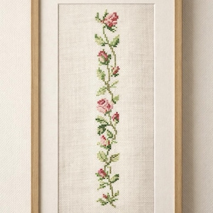 Può includere: Un'opera d'arte a punto croce incorniciata con un motivo floreale verticale. Mostra rose rosa e foglie verdi su uno sfondo color crema. La cornice è in legno chiaro. È un pezzo decorativo.