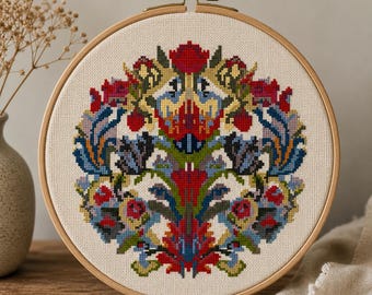 May include: A circular wooden embroidery hoop containing a cross-stitch pattern on off-white fabric. The design features a symmetrical, intricate floral and leaf motif in vibrant shades of red, blue, green, and yellow, centered on the fabric.