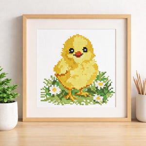 May include: A framed cross-stitch of a yellow chick with a red beak and black eyes, sitting on green grass with white daisies. The artwork is in a light brown wooden frame, displayed on a wooden surface.