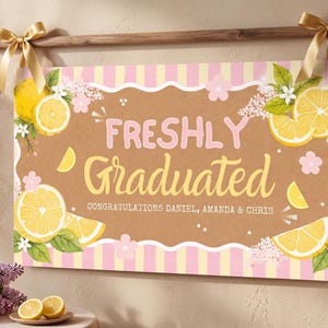 May include: A graduation banner with the words "FRESHLY GRADUATED" in pink and yellow, decorated with lemon slices and flowers. The banner is hanging on a wooden rod with gold bows. Balloons and flowers are in the background.