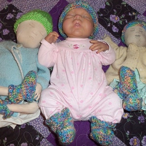May include: Three handmade cloth dolls wearing knitted hats and booties. The dolls are dressed in a blue sweater, a pink dress, and a cream cardigan. The dolls are lying on a purple and black patterned quilt.