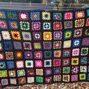 Pode incluir: Uma manta de crochê colorida feita de quadrados de avó em várias cores, incluindo rosa, azul, verde, amarelo e laranja. A manta está pendurada em um varal ao ar livre.