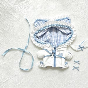 May include: A white hooded jacket with a blue and white plaid lining, featuring cat-ear shaped hood details, white ruffled trim, a blue zipper, and a blue harness strap. Includes a separate pair of small white wings and two tiny blue bows.