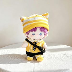 May include: A plush doll with purple hair wearing a yellow and white striped cat-ear beanie, a yellow sweater vest over a collared shirt, a black and white striped tie, black pants, yellow and white shoes, and a small black crossbody bag.