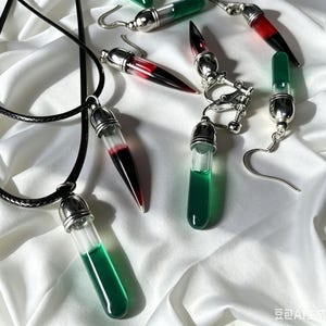 Könnte beinhalten: Eine Schmuckkollektion mit silberfarbenen Metallkappen und durchsichtigen Glasphiolen, die mit roter oder grüner Flüssigkeit gefüllt sind. Das Set enthält einen Anhänger an einer schwarzen Kordel, mehrere Ohrhänger und ein Paar Ohrclips.
