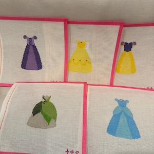 May include: Needlepoint canvases featuring colorful dress designs. The dresses are in shades of purple, yellow, green, and blue, each with unique details. The canvases have a white background with a pink border.