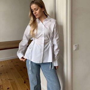 Op de afbeelding: Een witte blouse met lange mouwen en strikdetails in de taille, gecombineerd met lichtblauwe jeans. De foto is binnenshuis genomen, met natuurlijk licht dat de outfit belicht.