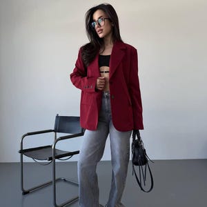 Puede incluir: Un blazer burdeos de diseño clásico, combinado con vaqueros grises y zapatillas negras. El conjunto incluye un top negro y un bolso negro. El fondo es una pared blanca lisa y un suelo gris.