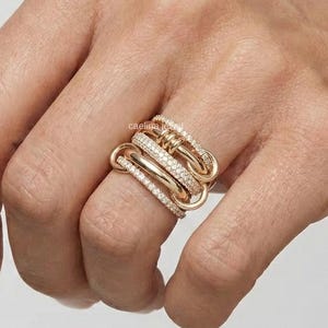 Könnte beinhalten: Ein goldfarbener Ring mit mehreren ineinandergreifenden Bändern, von denen einige mit kleinen, funkelnden Edelsteinen besetzt sind. Der Ring wird auf einer Hand vor neutralem Hintergrund getragen und zeigt sein aufwendiges Design.