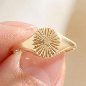 Könnte beinhalten: Ein goldfarbener Ring mit einem Sonnenstrahlen-Design. Der Ring wird zwischen Fingern gehalten, was seine polierte Oberfläche und die filigrane Detailarbeit zeigt. Das Design des Rings zeigt einen zentralen Kreis mit radialen Linien, die ein sonnenähnliches Muster bilden. Der Ring ist ein Schmuckstück.