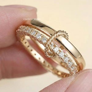 Könnte beinhalten: Ein goldfarbenes Ring-Set mit Knoten-Design. Ein Band ist mit kleinen, klaren Edelsteinen besetzt, das andere ist ein glattes, poliertes Band. Das Knotendetail verleiht dem Design des Rings eine einzigartige Note.