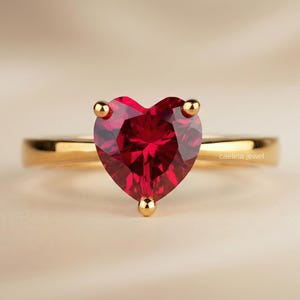 Könnte beinhalten: Ein goldener Ring mit einem herzförmigen, facettierten, tiefroten Edelstein. Der Edelstein wird von drei goldenen Krappen gehalten. Der Ring ist aus poliertem Gold. Der Text "caelina jewel" ist auf dem Ring sichtbar.