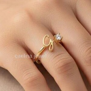 Puede incluir: Un anillo dorado con la letra "G" en cursiva y una gema transparente. El anillo está en un dedo. El texto "caelina jewel" es visible.