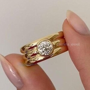 Könnte beinhalten: Ein goldener Ring mit einem großen, runden, klaren Edelstein in der Mitte. Der Ring hat ein einzigartiges, geschichtetes Design mit strukturierter Oberfläche. Der Text "caelina jewel" ist auf dem Ring zu sehen.
