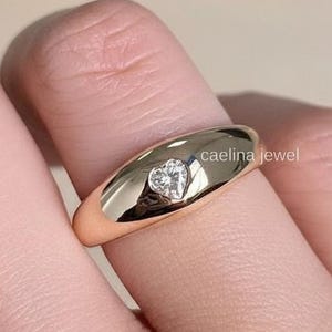Puede incluir: Un anillo de oro con un diamante en forma de corazón engastado en el centro. El anillo es liso y pulido, con el texto "caelina jewel" en el lateral. El anillo está en un dedo.