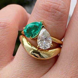 May include: Three gold rings with gemstones. One ring features a pear-shaped, emerald-green gemstone. Another ring has a pear-shaped, clear gemstone. The third ring is a thick, curved gold band. The rings are stacked on a finger.