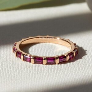 May include: A rose gold ring with a continuous band of rectangular, deep red gemstones. The ring is set against a white surface with a green leaf and a white stone in the background. The ring is a piece of jewelry.