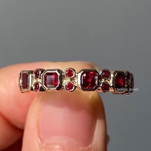 Könnte beinhalten: Ein goldfarbener Ring mit abwechselnd quadratischen und runden roten Edelsteinen, die um das Band herum angeordnet sind. Der Ring wird von den Fingern einer Person gehalten. Der Text "caelina jewel" ist auf dem Ring sichtbar.