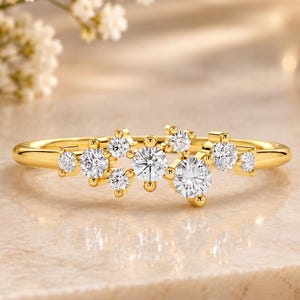 Könnte beinhalten: Ein Goldring mit einer Ansammlung mehrerer runder, klarer, facettierter Steine, die in einem verstreuten, asymmetrischen Muster auf der Oberseite des Rings angeordnet sind. Der Metallring ist poliert und glatt und hat ein helles, glänzendes Finish.