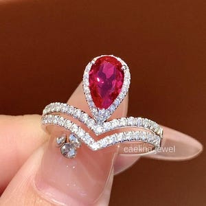 Könnte beinhalten: Ein silberner Ring mit einem tropfenförmigen, leuchtend roten Edelstein, umgeben von kleinen weißen Steinen. Der Ring hat ein Doppelband-Design in V-Form. Der Text "caelina jewel" ist sichtbar.