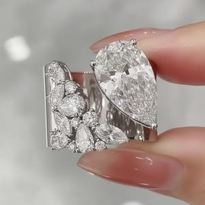 Könnte beinhalten: Ein schimmernder Silberring mit einem großen birnenförmigen Diamanten und einer Ansammlung kleinerer Diamanten. Das Design des Rings umfasst ein rechteckiges Band mit einer blütenartigen Anordnung von Diamanten, wodurch ein luxuriöses und auffälliges Schmuckstück entsteht.