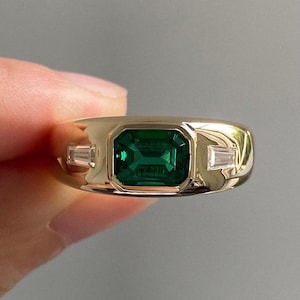 Könnte beinhalten: Ein goldener Ring mit einem großen, smaragdgeschliffenen grünen Edelstein in der Mitte, flankiert von zwei kleineren, rechteckigen, klaren Edelsteinen. Der Ring hat eine polierte, reflektierende Oberfläche.