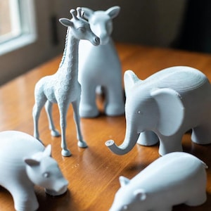 May include: A collection of light gray animal figurines, including a giraffe, elephant, and hippos, are arranged on a wooden surface. The giraffe stands tall, while the elephant has a curled trunk. The figurines are detailed and have a smooth finish.