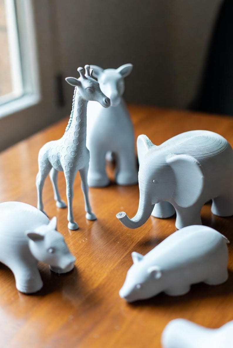 May include: A collection of light gray animal figurines, including a giraffe, elephant, and hippos, are arranged on a wooden surface. The giraffe stands tall, while the elephant has a curled trunk. The figurines are detailed and have a smooth finish.