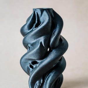 May include: A dark gray, abstract vase with a twisted, organic design. The vase has a textured surface and a narrow opening at the top. The design features several openings along the sides.