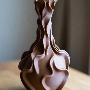 May include: A brown, decorative vase with a unique, flowing design. The vase has a ruffled top and a textured, wave-like pattern throughout its body. The vase is made of a solid material and sits on a wooden surface.