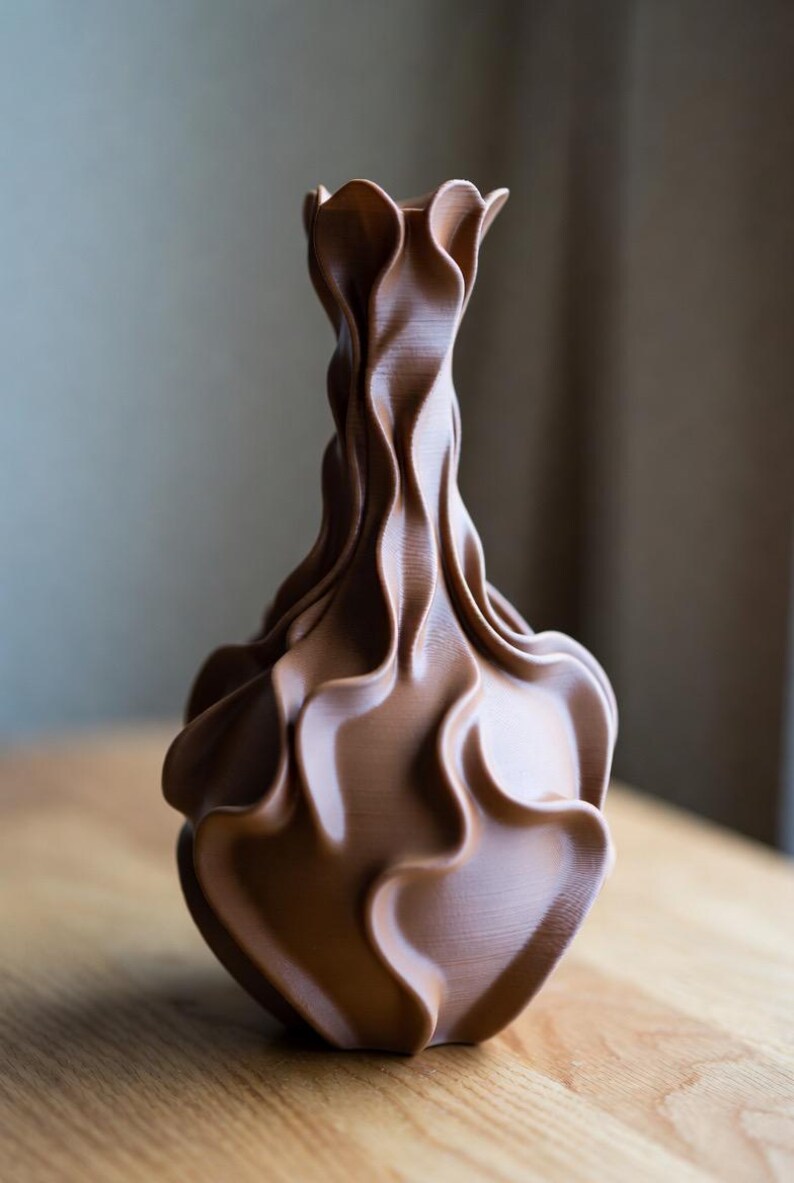 May include: A brown, decorative vase with a unique, flowing design. The vase has a ruffled top and a textured, wave-like pattern throughout its body. The vase is made of a solid material and sits on a wooden surface.