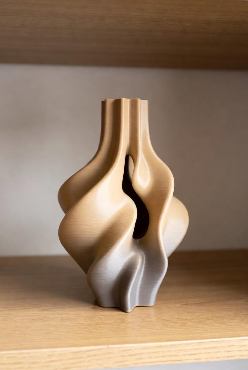 May include: A beige, abstract vase with a unique, flowing design. The vase has a textured surface and a dark interior visible through a central opening. The form is reminiscent of organic shapes, suitable for home decor.