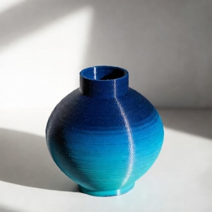 May include: A small, round vase with a gradient of blue hues, transitioning from a deep navy at the top to a vibrant turquoise at the base. The vase has a cylindrical neck and a textured surface, perfect for displaying flowers or as a decorative piece.