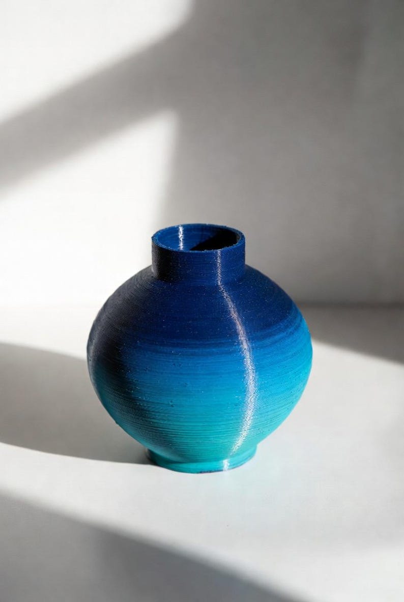 May include: A small, round vase with a gradient of blue hues, transitioning from a deep navy at the top to a vibrant turquoise at the base. The vase has a cylindrical neck and a textured surface, perfect for displaying flowers or as a decorative piece.