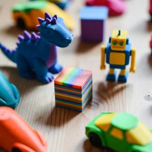 May include: A collection of colorful 3D-printed toys, including a blue dinosaur with purple accents, a yellow and blue robot, and various toy cars in orange and green. A small, square block with rainbow-colored layers is also present.