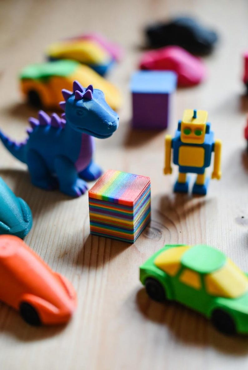 May include: A collection of colorful 3D-printed toys, including a blue dinosaur with purple accents, a yellow and blue robot, and various toy cars in orange and green. A small, square block with rainbow-colored layers is also present.
