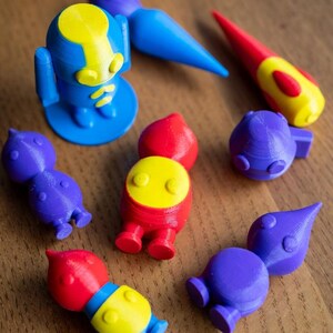 May include: A collection of colorful 3D-printed toys in blue, red, yellow, and purple. The toys include robot-like figures, a paintbrush, and other abstract shapes. The toys are arranged on a wooden surface.