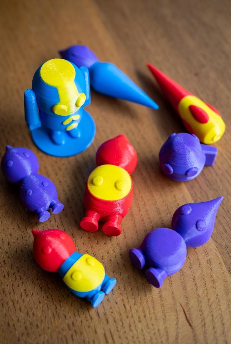 May include: A collection of colorful 3D-printed toys in blue, red, yellow, and purple. The toys include robot-like figures, a paintbrush, and other abstract shapes. The toys are arranged on a wooden surface.