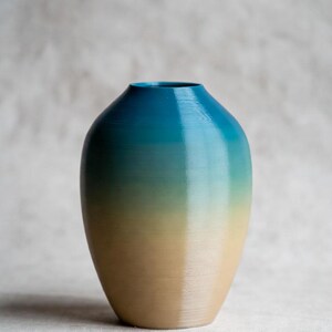 May include: A decorative vase with a gradient color scheme. The top of the vase is a deep teal, transitioning to a light green, and finally to a tan base. The vase has a rounded shape and a smooth finish.