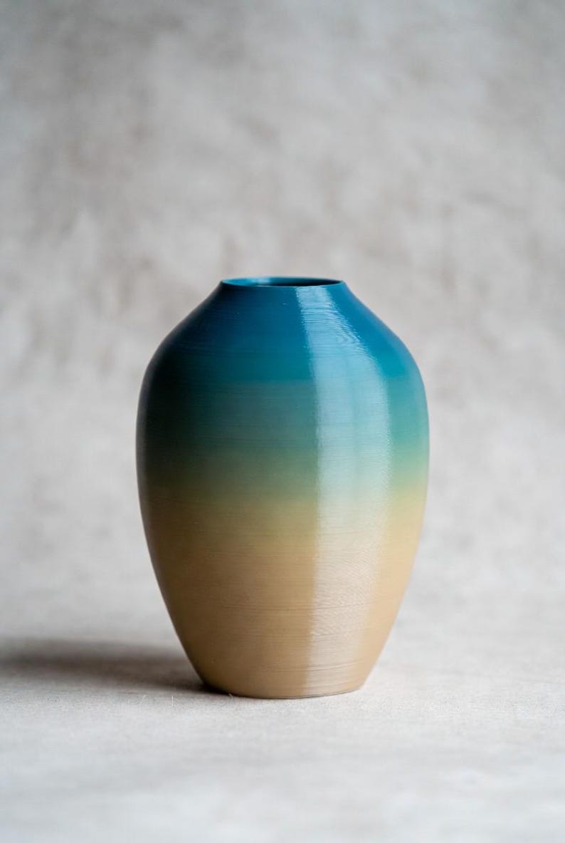 May include: A decorative vase with a gradient color scheme. The top of the vase is a deep teal, transitioning to a light green, and finally to a tan base. The vase has a rounded shape and a smooth finish.