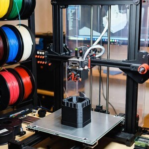 May include: A 3D printer in operation, constructing a black, multi-tiered structure. The printer has a black frame with orange accents, a clear enclosure, and a heated bed. Spools of colorful filament are visible on a rack. The text "3D PRINTING SERVICES" is in the background.