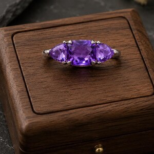 Könnte beinhalten: Ein silberner Ring mit drei großen, leuchtend violetten Edelsteinen, präsentiert in einer dunklen Holzschatulle. Die Edelsteine des Rings sind dreieckig und in einem silbernen Band gefasst. Die Schatulle ist dunkelbraun mit einem kleinen goldfarbenen Verschluss.