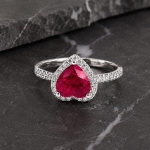 Könnte beinhalten: Ein silberfarbener Ring mit einem herzförmigen roten Edelstein in der Mitte, der von einem Kranz aus kleinen weißen Steinen umgeben ist. Die Ringschiene ist ebenfalls mit kleinen weißen Steinen besetzt, was für ein Funkeln sorgt.